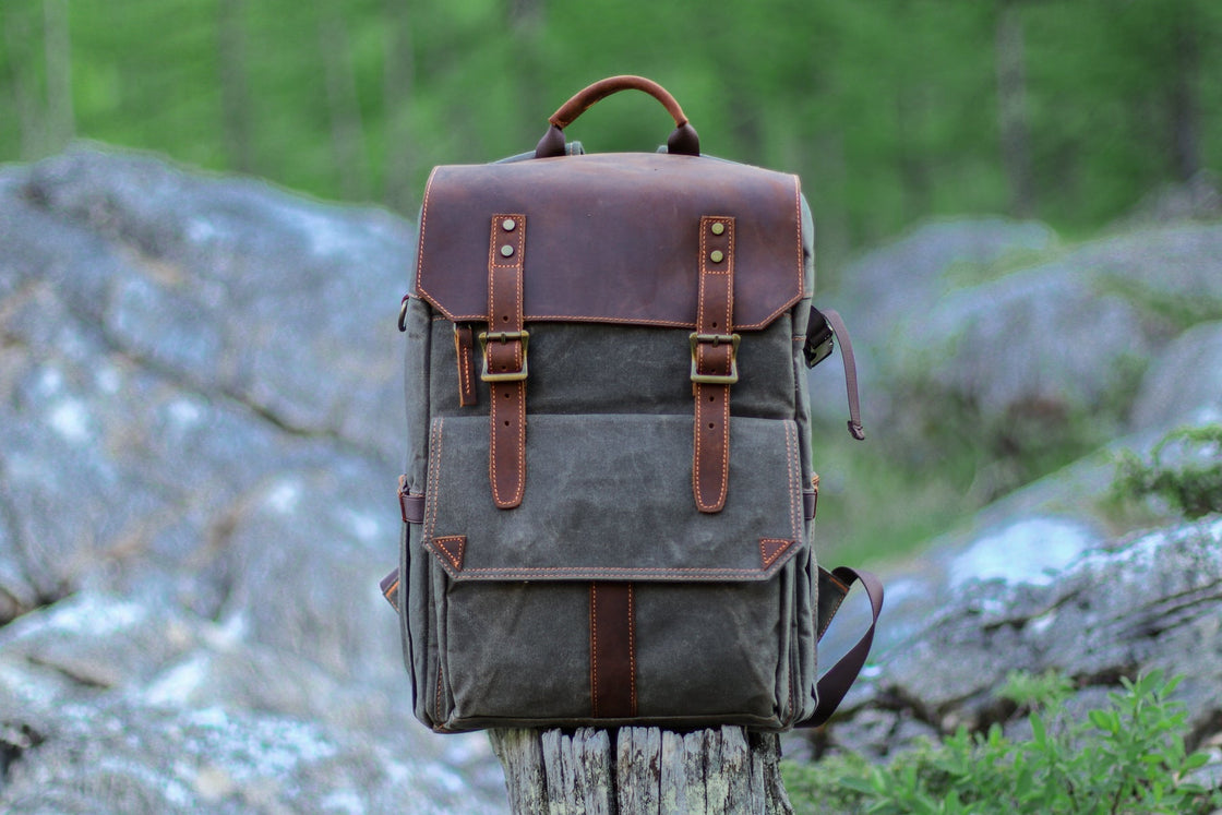 Dark grey-green Algonquin waxed canvas camera backpack standing on a tree stump with alpine rocks behind, leather flap and orange contrast stitching