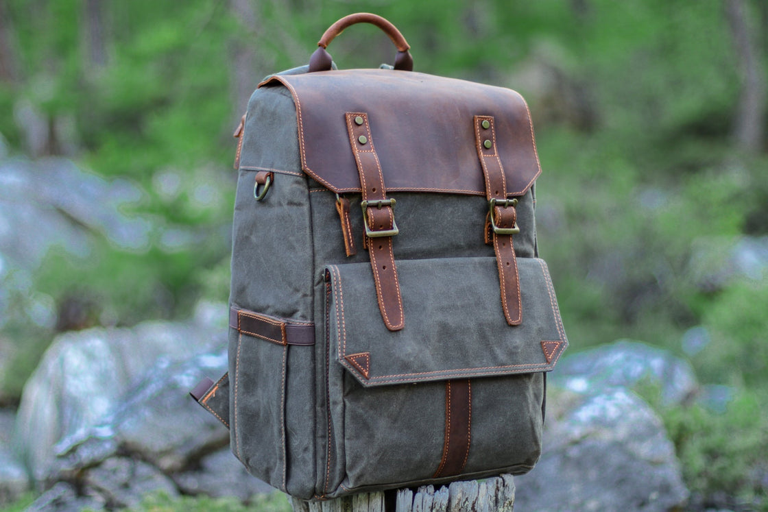 Three-quarter view of the dark grey-green Algonquin on a wooden stump, with leather flap, side bottle pocket and orange topstitching catching daylight