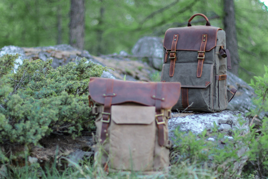 Two Algonquin DSLR camera backpacks staged in an alpine pine forest, khaki at the base and dark grey-green on a granite boulder behind, comparing the colorways
