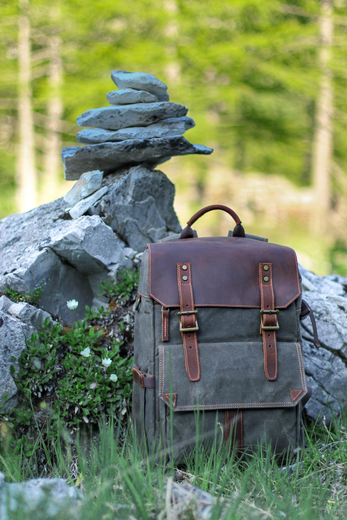 Sac à dos appareil photo Algonquin vert gris foncé mis en scène à côté d'un cairn de pierres dans une forêt de pins alpine, rabat en cuir et poche utilitaire visibles