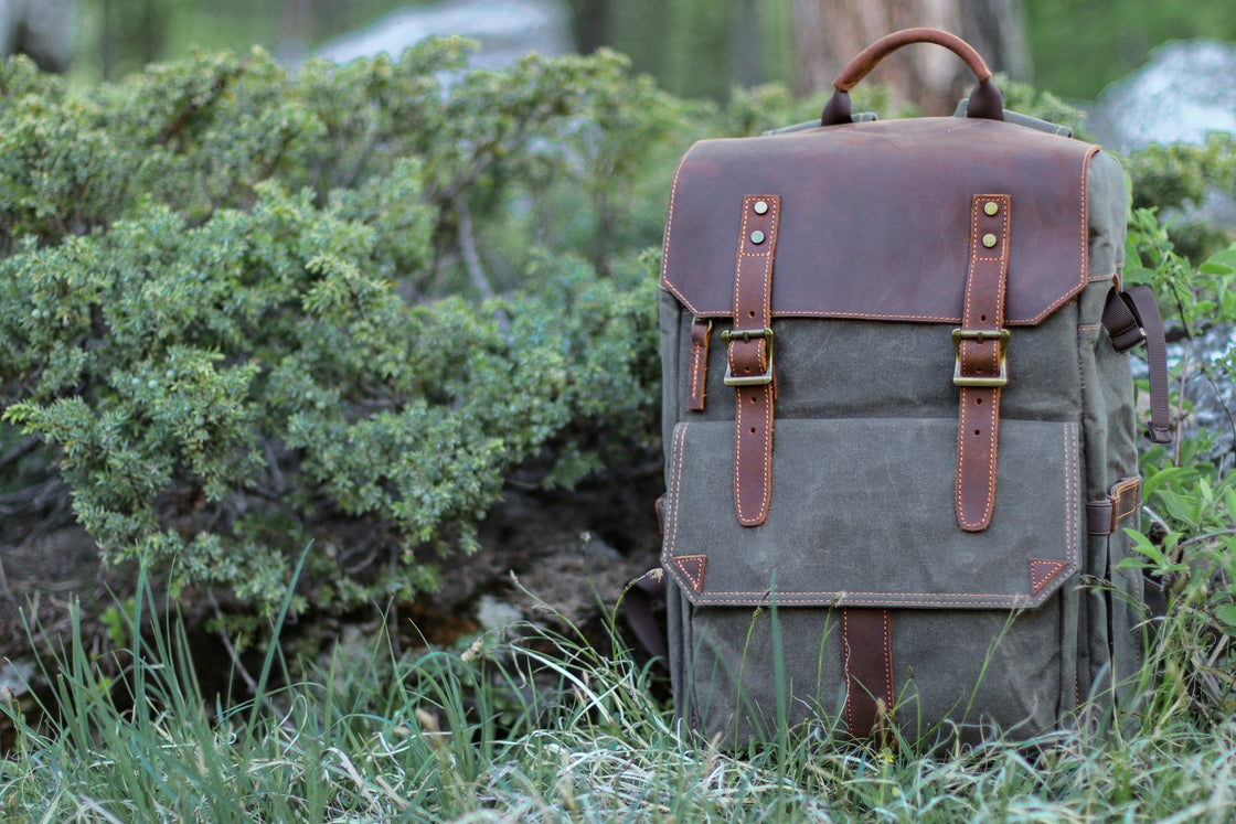 Sac à dos photographe Algonquin vert gris foncé posé sur l'herbe près de feuillage de genévrier, rabat en cuir et doubles fermetures à sangle avec surpiqûres orange