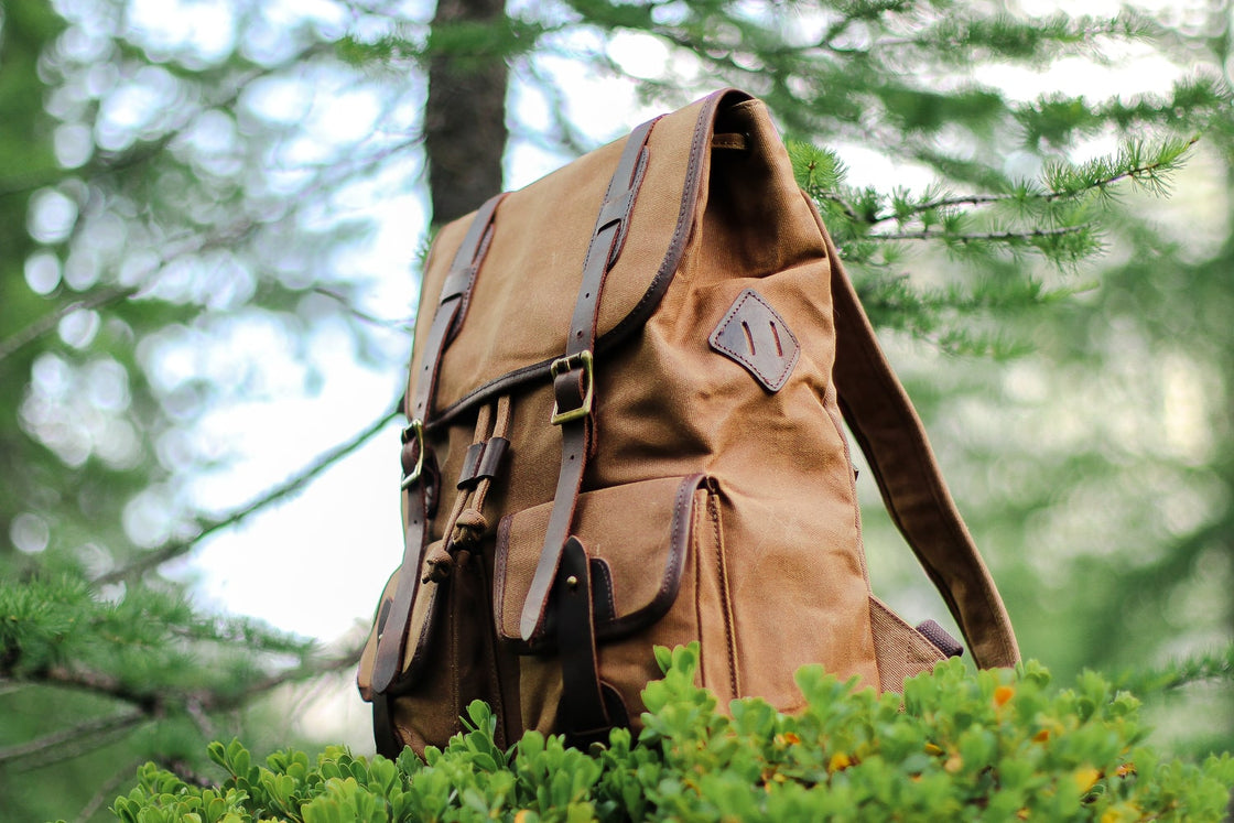 Three-quarter low-angle view of the khaki Calgary canvas utility backpack against alpine pine forest, with leather strap closures and dual cargo pockets in view