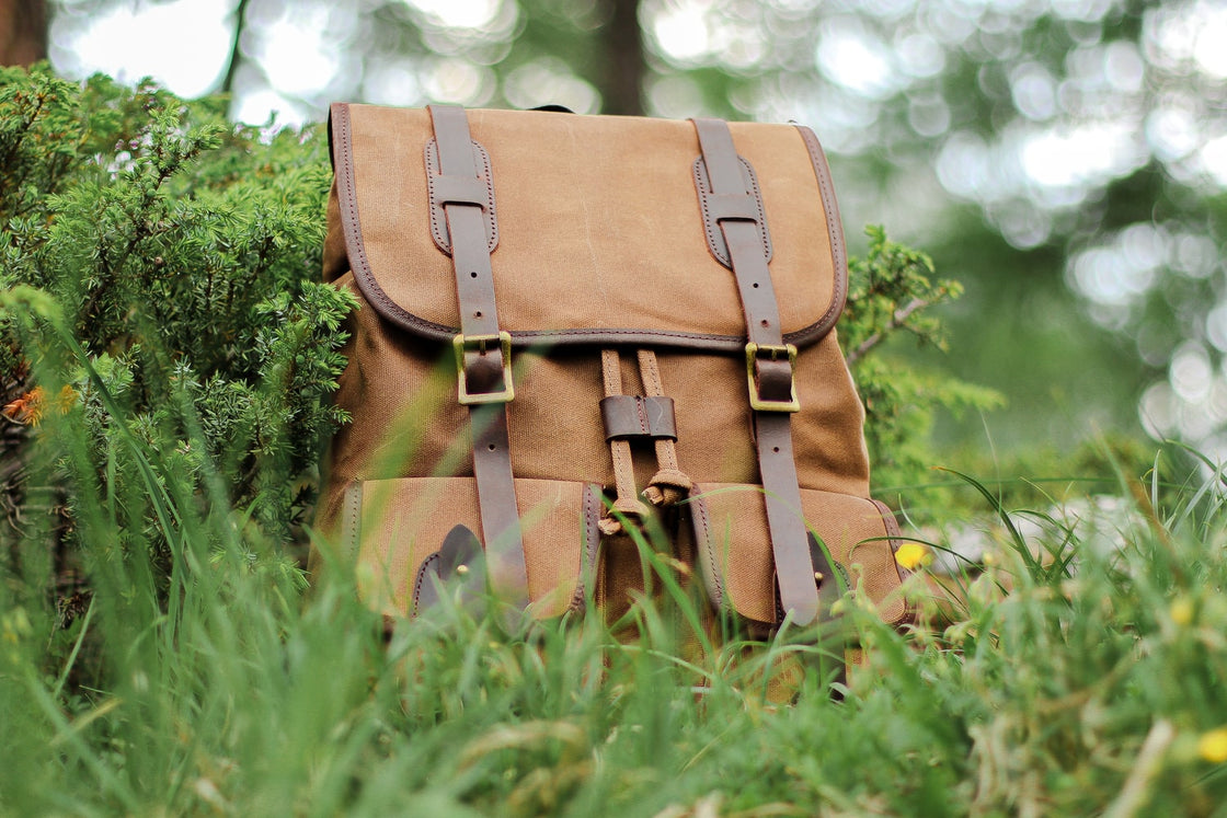 Three-quarter view of the khaki Calgary canvas backpack in tall alpine grass, with leather strap closures and dual front cargo pockets in soft daylight