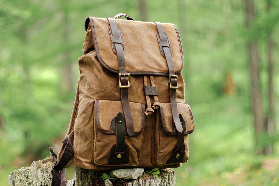 Khaki Calgary canvas rucksack standing on a wooden tree stump in a pine forest, with leather flap, drawstring closure and twin cargo pockets in view