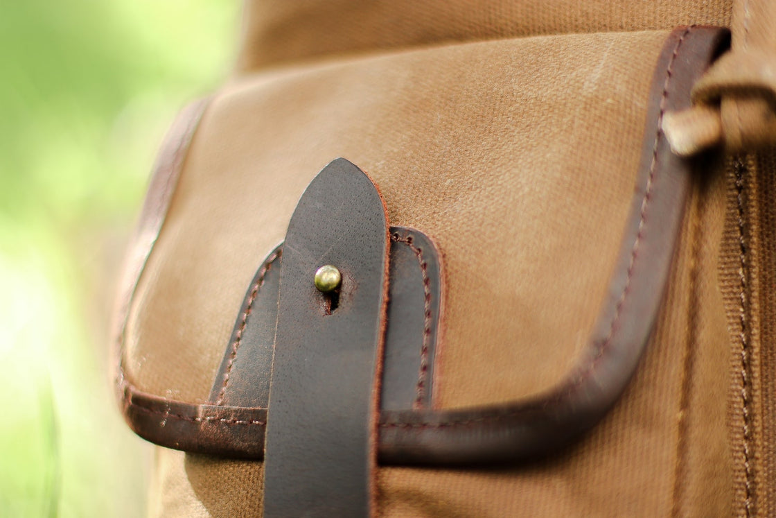 Close-up of the front cargo pocket flap with dark leather closure tab and brass snap on the khaki Calgary backpack