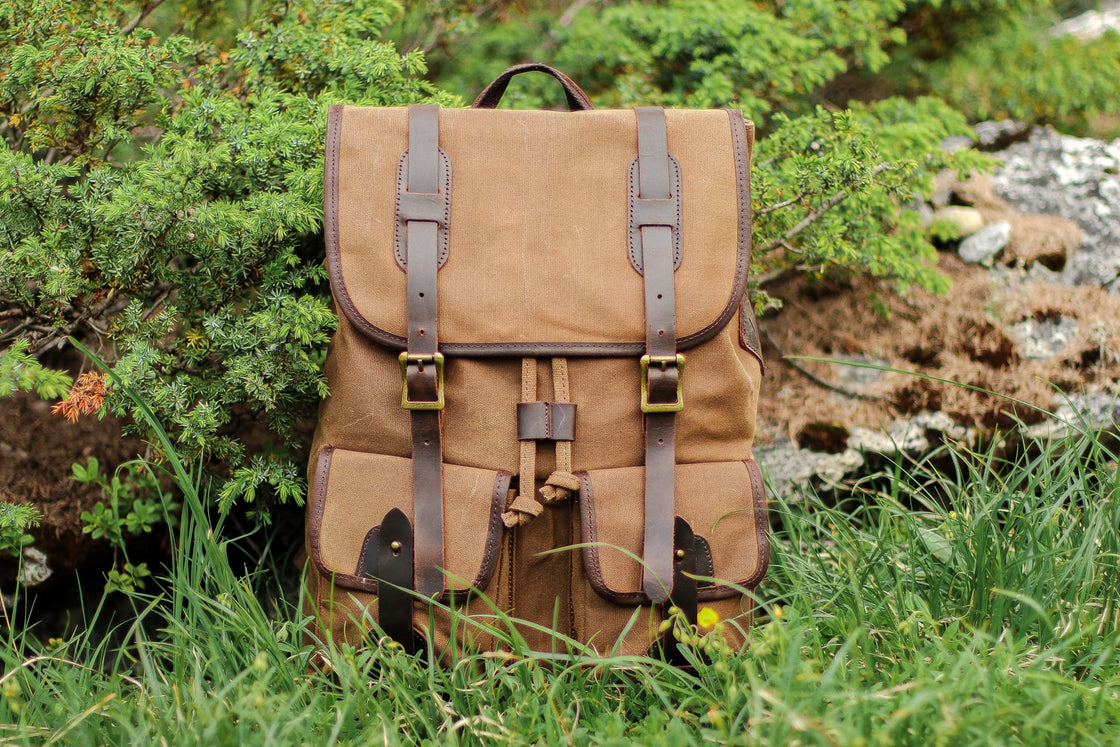 Vue de face du sac à dos voyage Calgary en toile cirée kaki sur l'herbe alpine près de feuillage de genévrier, rabat en cuir et doubles poches cargo avant visibles