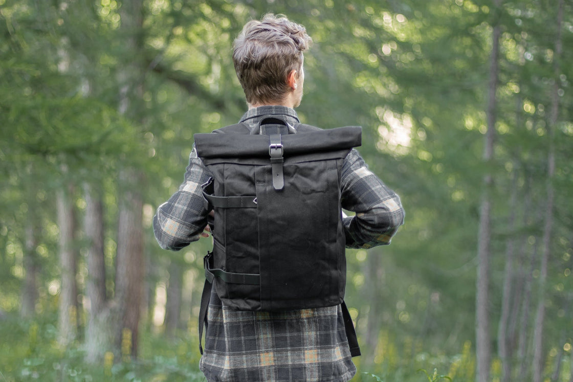 Homme portant le Gstaad noir vu de dos en forêt avec chemise plaid, sac roll-top imperméable