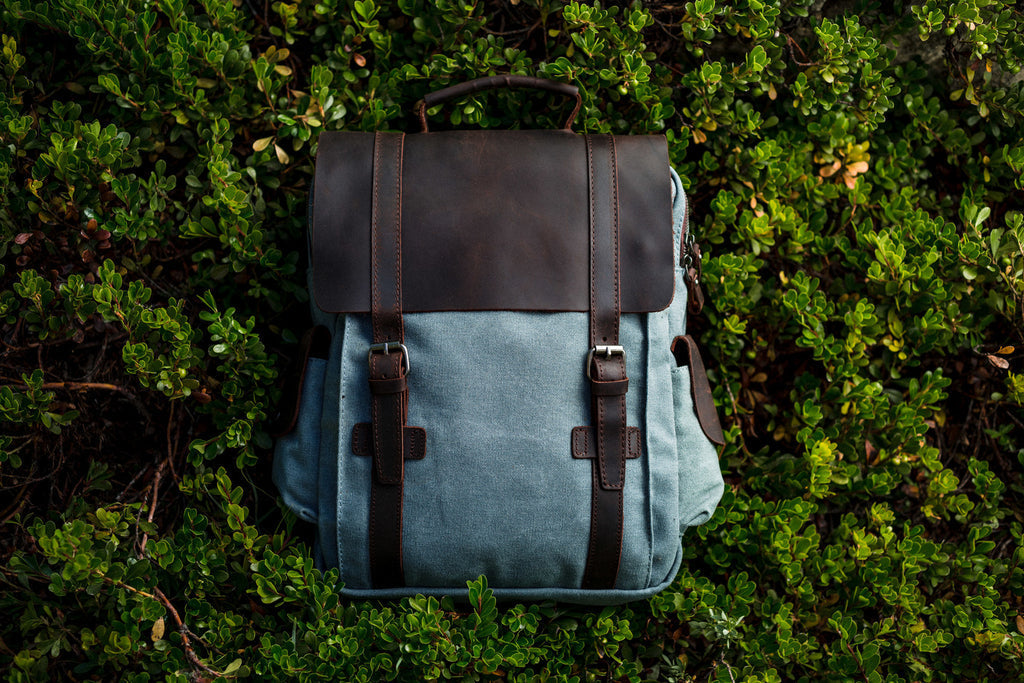 Top-down flat-lay of the light blue Manitoba small canvas backpack on a bilberry bush, with dark brown leather flap and twin strap closures