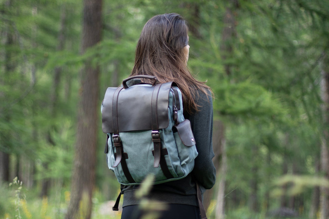 Woman wearing the light blue Manitoba small canvas backpack on her back in a pine forest, with leather flap and twin shoulder straps visible