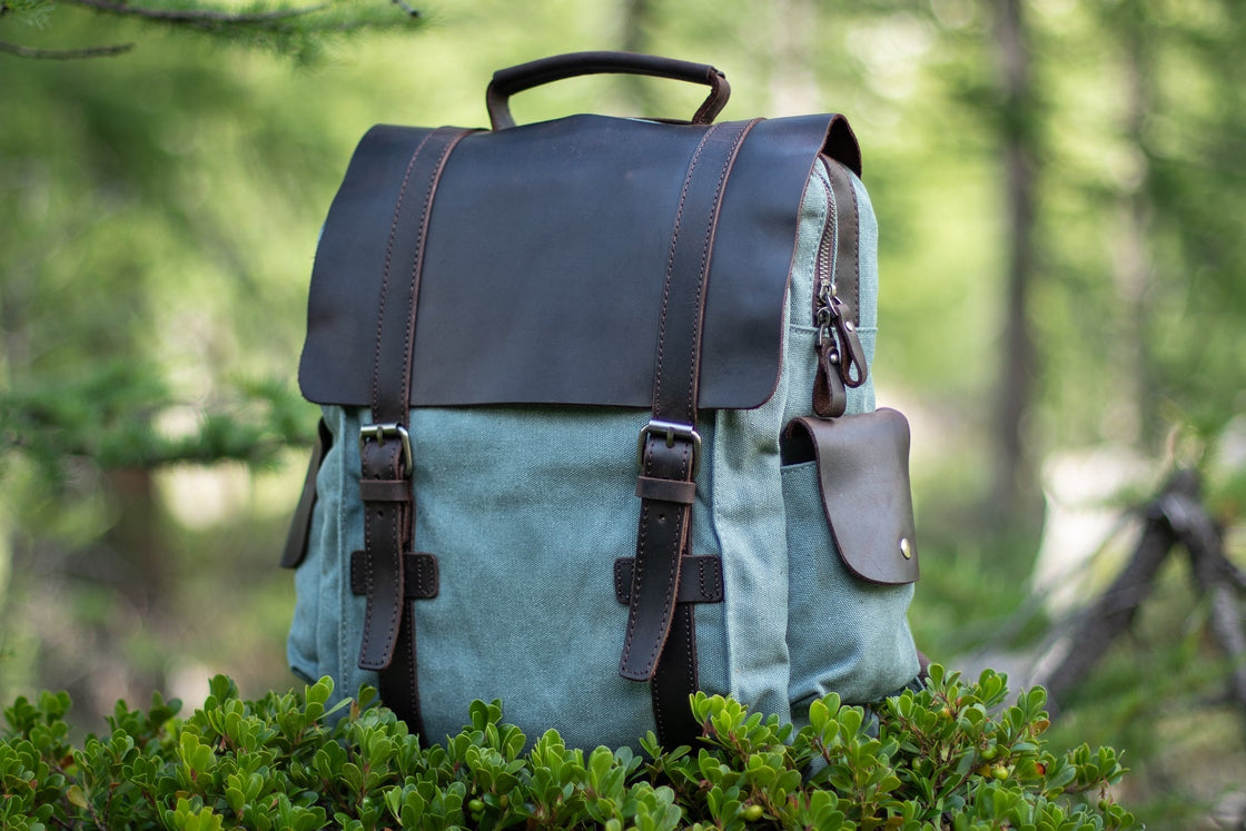 Three-quarter view of the light blue Manitoba small canvas backpack on a bilberry bush, with side leather pocket, brass snap and dark leather flap