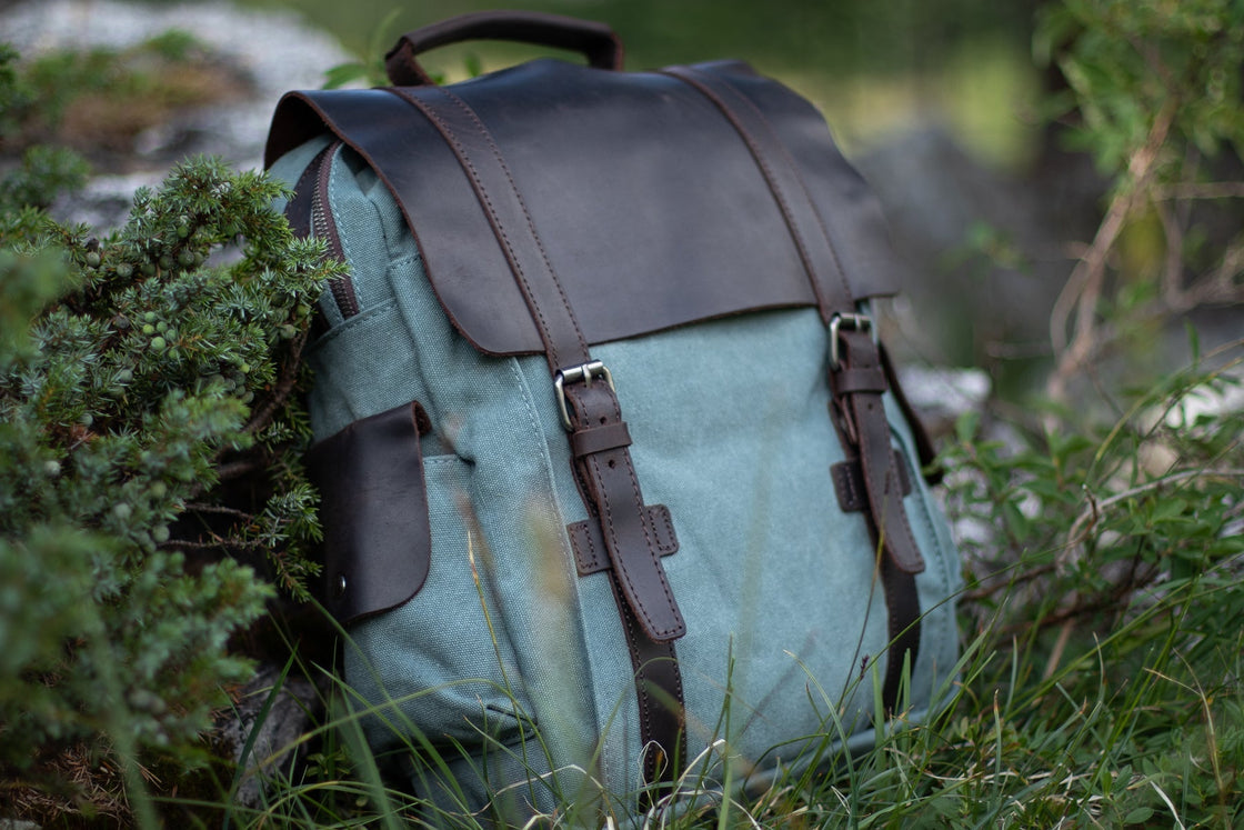 Three-quarter view of the light blue Manitoba small canvas backpack nestled in alpine grass and juniper, with leather flap and side cargo pocket