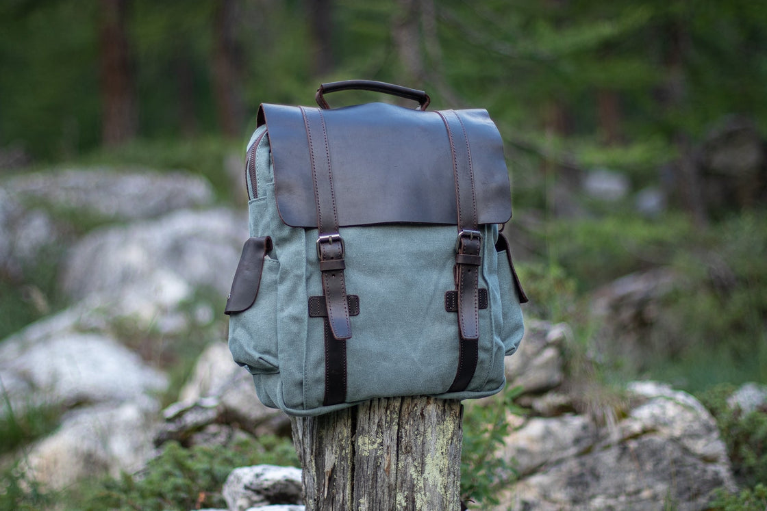Three-quarter view of the light blue Manitoba small canvas backpack on a tree stump in an alpine pine forest, with leather flap and side cargo pocket