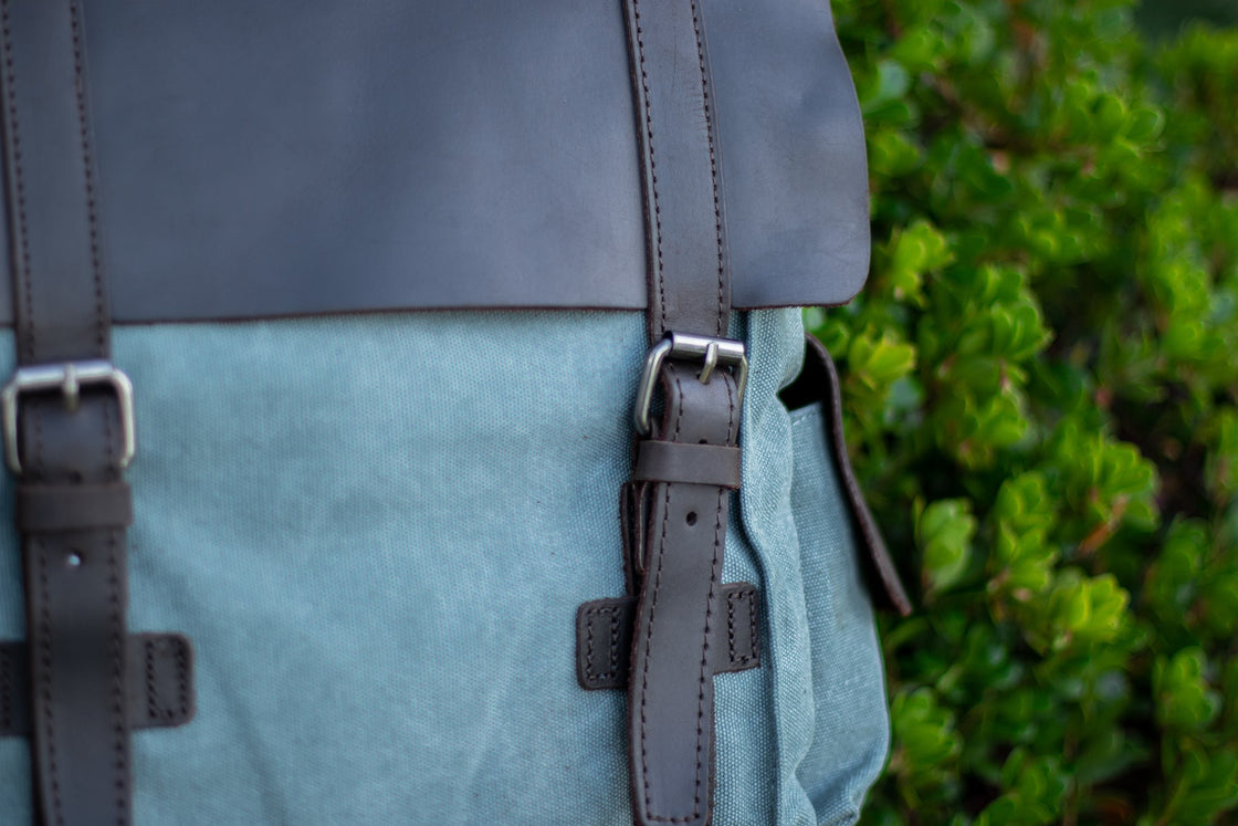 Vue trois-quarts du petit sac à dos femme Manitoba bleu clair niché dans l'herbe alpine et le genévrier, rabat en cuir et poche cargo visibles sous un angle doux