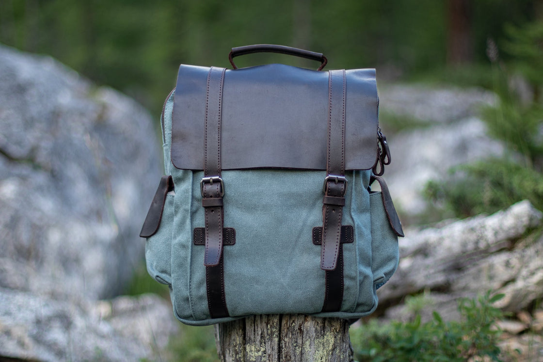 Vue trois-quarts du petit sac à dos Manitoba bleu clair sur une souche d'arbre dans une forêt de pins alpine, rabat en cuir et détail de la poche cargo