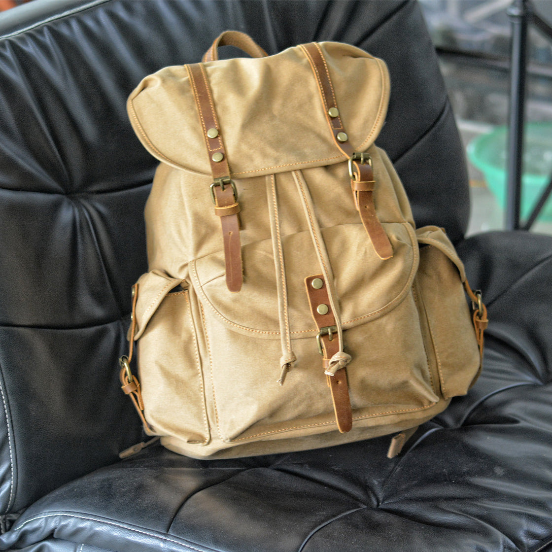Sac à dos Montreal en toile kaki posé sur un fauteuil lounge en cuir noir dans un showroom intérieur, rabat surdimensionné en cuir et poches latérales cargo visibles