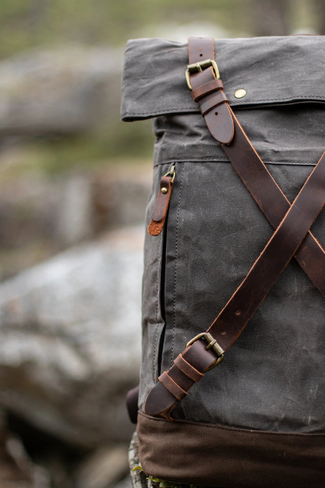 Vertical detail of the criss-cross full-grain leather straps on the dark grey Oslo waxed canvas rucksack, brass buckles and side zip pocket pull visible