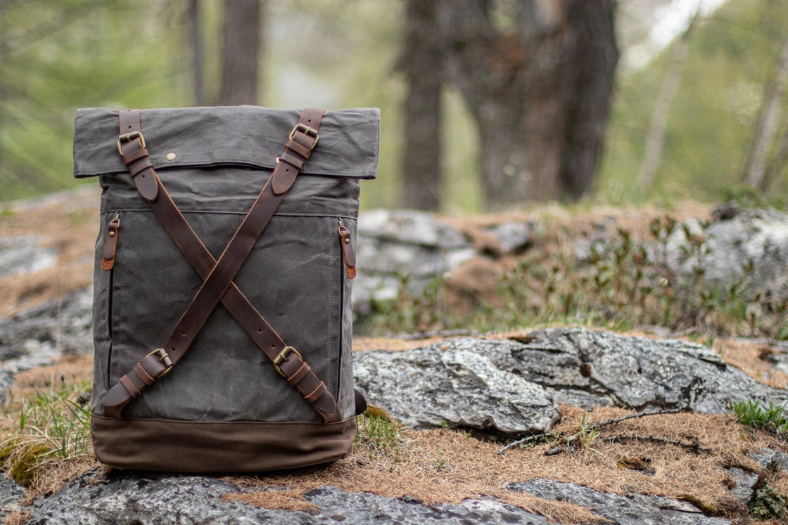 Dark grey Oslo waxed canvas rucksack standing on a moss-covered alpine rock in a pine forest, criss-cross leather strap closure and brown canvas base