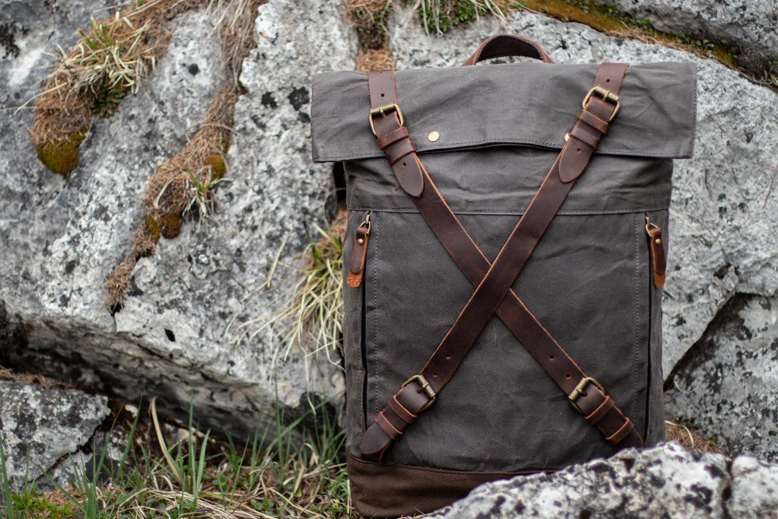 Sac à dos en toile Oslo en toile cirée gris foncé reposant sur des rochers de granit alpins, fermetures à sangles entrecroisées en cuir et boucles à rouleau en laiton captant la lumière du jour