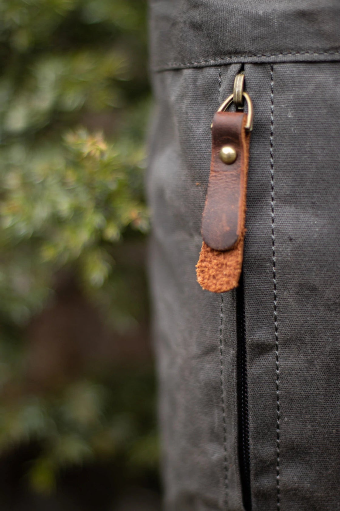 Gros plan de la tirette de fermeture éclair YKK latérale avec languette en cuir pleine fleur et D-ring en laiton vieilli sur le sac à dos en toile Oslo gris foncé