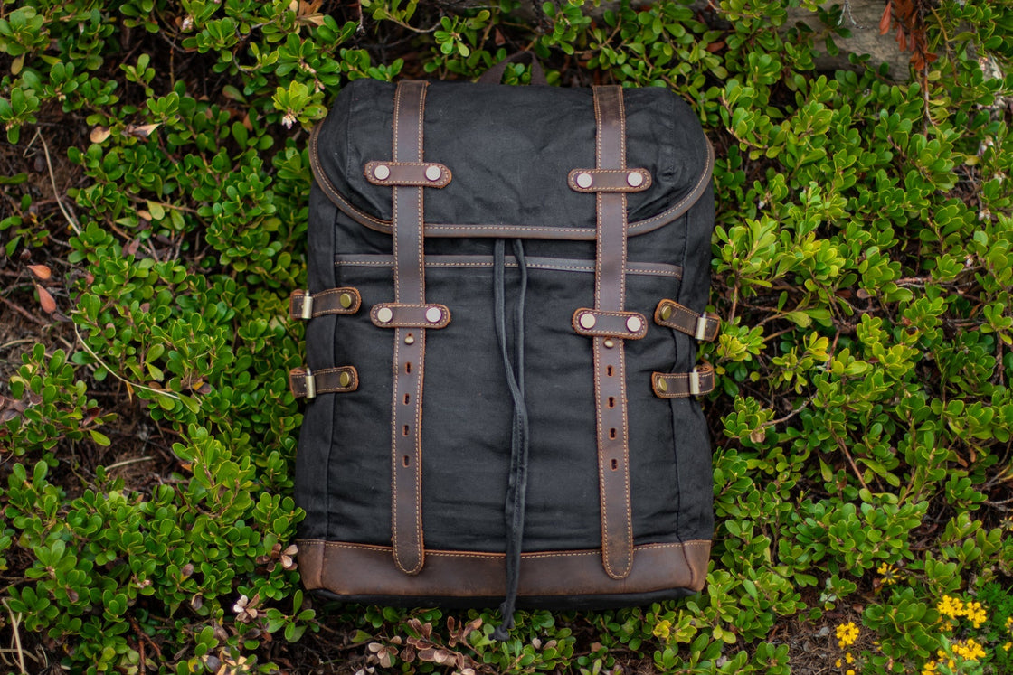 Sac à dos style vintage en toile cirée noire avec sangles cuir crazy horse, boucles laiton et poches latérales, photographié de dessus sur des buissons forestiers, DIYION Rostock