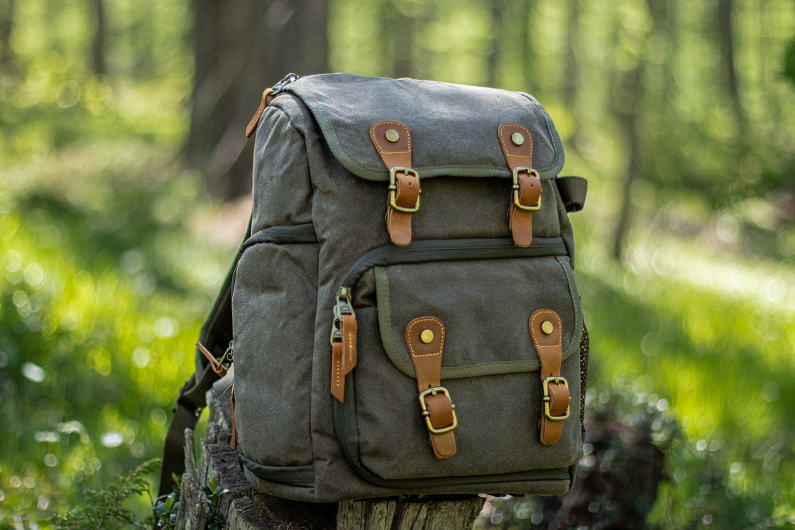 Army green Vilnius leather and canvas camera backpack on a tree stump in the forest with twin leather strap closures