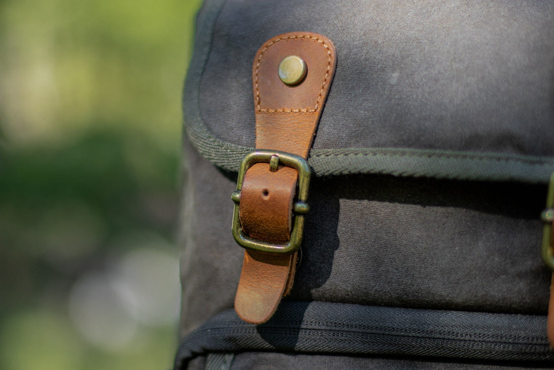 Macro detail of an antiqued brass buckle and Crazy Horse leather strap on the Vilnius army green canvas camera backpack