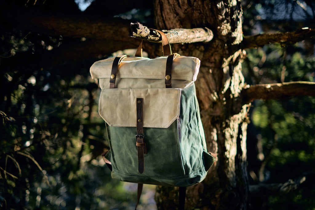 Front view of the beige Yukon vintage canvas backpack on a forest floor, leather flap and side cargo pockets visible