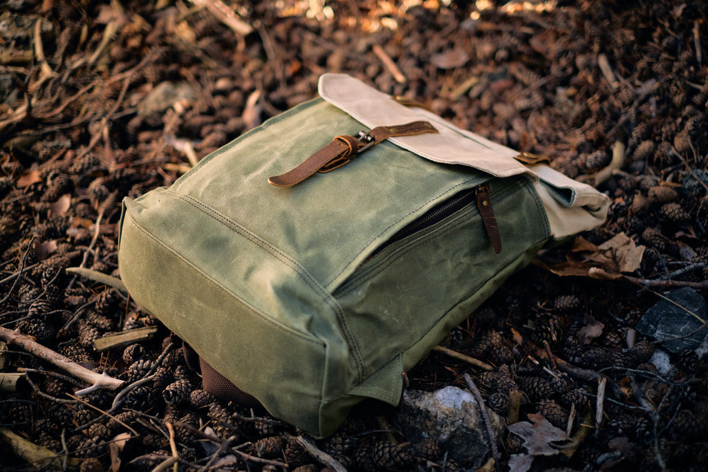 Beige Yukon vintage canvas rucksack in alpine grass beside juniper, leather flap and brass buckles catching daylight